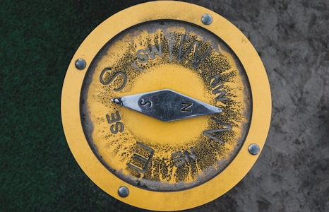 A yellow circular compass mounted on a flat surface, showing cardinal and intercardinal directions. The compass has visible wear, with some black marks surrounding the directional letters. The needle is pointing towards the north. There are four bolts securing the compass.