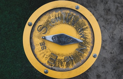 A yellow circular compass mounted on a flat surface, showing cardinal and intercardinal directions. The compass has visible wear, with some black marks surrounding the directional letters. The needle is pointing towards the north. There are four bolts securing the compass.