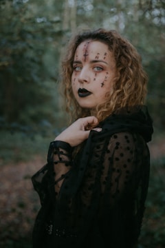 Close-up of a model with intricate dark fantasy makeup blending deep greens and blacks, surrounded by misty woodland.