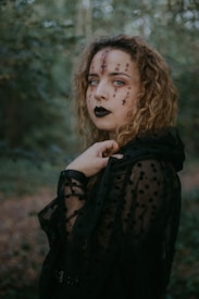A woman with curly hair and dark makeup stands in a forest. Her face is decorated with black symbols and markings, and she wears a black sheer, polka-dotted cloak. The forest background is lush and green, creating a mystical atmosphere.