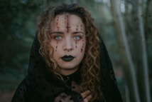 A woman with curly hair and striking blue eyes is adorned with black makeup and intricate symbols drawn across her face. She wears a black veil or cloak and stands amidst a forest setting. Her expression is intense and her hand is partially visible near her chest.