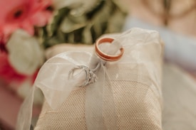 Two rings, one gold and one silver with a diamond, rest atop a textured cream cushion or fabric, tied together with a sheer white ribbon. In the background, there are blurred flowers in shades of red and cream, adding a romantic atmosphere.