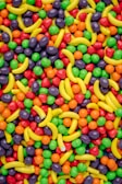 A vibrant assortment of colorful candies, including banana-shaped candies and small round sweets in red, orange, green, and purple hues. The candies are randomly scattered, creating a visually striking and cheerful pattern.