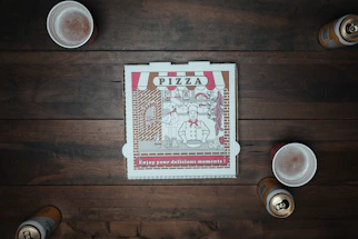 A closed pizza box with a vintage design sits at the center of a wooden table surrounded by three foamy drinks and two open cans. The box features a chef illustration alongside pizza-themed decorations.