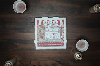 A closed pizza box with a vintage design sits at the center of a wooden table surrounded by three foamy drinks and two open cans. The box features a chef illustration alongside pizza-themed decorations.