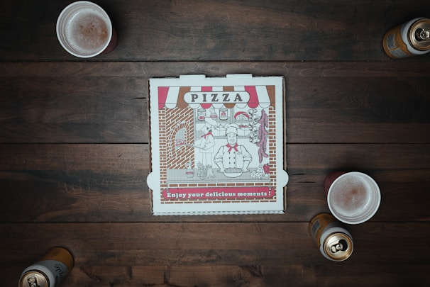 A closed pizza box with a vintage design sits at the center of a wooden table surrounded by three foamy drinks and two open cans. The box features a chef illustration alongside pizza-themed decorations.