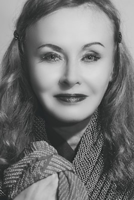 A woman with wavy hair smiles softly at the camera. She wears a patterned garment and has styled hair with slight waves. The image is in black and white, emphasizing the textures and contrasts on her face and clothing.