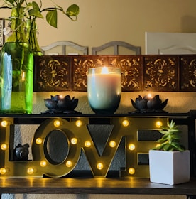 A decorative arrangement featuring a shelf with illuminated letters spelling 'LOVE' surrounded by small lights. On top of the shelf, there is a lit candle in a blue-green jar, flanked by two dark lotus-shaped candle holders. A green plant in a transparent bottle and a small succulent in a white pot add a touch of nature to the scene. The background includes a decorative wooden panel with ornate cut-out designs.
