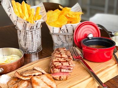 A wooden board holds a piece of grilled steak with grill marks, accompanied by slices of cooked meatloaf. Beside it is a vibrant red small pot filled with sauce, and a fork and knife rest nearby. In metal baskets lined with newspaper-style paper are French fries and waffle-cut chips. A small copper pan contains seasoned fried eggs.