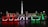 Illuminated letters spelling 'BUDAPEST' with each letter glowing in colors of red, white, and green against a nighttime backdrop. Behind the letters, a monument with statues and a tall pillar topped with a statue are visible, likely indicating a city square setting.