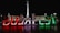 Illuminated letters spelling 'BUDAPEST' with each letter glowing in colors of red, white, and green against a nighttime backdrop. Behind the letters, a monument with statues and a tall pillar topped with a statue are visible, likely indicating a city square setting.