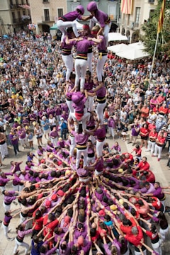 A large crowd gathers in a town square to watch a human tower being built. People are dressed in matching purple and white outfits, with helmets for safety. The human tower consists of several tiers, with participants standing on each other's shoulders. Bystanders surround the tower, observing and taking pictures.