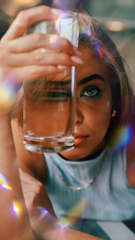 A person is holding a glass of water close to their face with one eye visible through the glass. Their expression seems contemplative, and they are surrounded by refracted light creating colorful reflections.