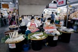 Three women at a busy market are seated behind tables filled with various goods, including wrapped items on trays covered with banana leaves. The setting is bustling with a variety of shops displaying products and signs in a language that suggests a Southeast Asian location. The women appear relaxed as they either work or engage with their surroundings.