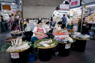 Three women at a busy market are seated behind tables filled with various goods, including wrapped items on trays covered with banana leaves. The setting is bustling with a variety of shops displaying products and signs in a language that suggests a Southeast Asian location. The women appear relaxed as they either work or engage with their surroundings.