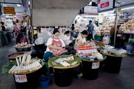 Three women at a busy market are seated behind tables filled with various goods, including wrapped items on trays covered with banana leaves. The setting is bustling with a variety of shops displaying products and signs in a language that suggests a Southeast Asian location. The women appear relaxed as they either work or engage with their surroundings.