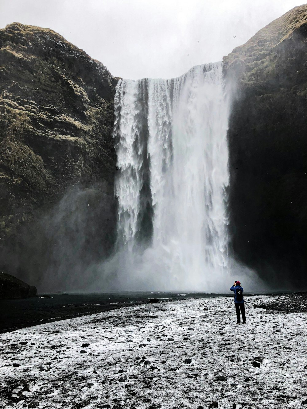 homme en veste bleue debout près des chutes d’eau pendant la journée