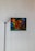 A modern art painting with abstract faces in warm hues of orange, red, and yellow is mounted on a white wall. The background contains shades of blue and green, creating a striking contrast. A black floor lamp with a white lampshade stands to the left of the painting, contributing to the minimalist decor.