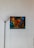 A modern art painting with abstract faces in warm hues of orange, red, and yellow is mounted on a white wall. The background contains shades of blue and green, creating a striking contrast. A black floor lamp with a white lampshade stands to the left of the painting, contributing to the minimalist decor.