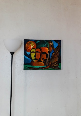 A modern art painting with abstract faces in warm hues of orange, red, and yellow is mounted on a white wall. The background contains shades of blue and green, creating a striking contrast. A black floor lamp with a white lampshade stands to the left of the painting, contributing to the minimalist decor.