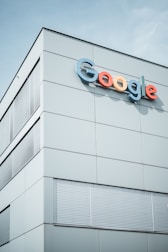 A modern building with the Google logo displayed prominently on the upper section. The structure features minimalist design with large, smooth panels and multiple sets of window blinds. The sky is clear in the background.