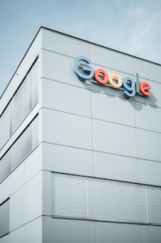 A modern building with the Google logo displayed prominently on the upper section. The structure features minimalist design with large, smooth panels and multiple sets of window blinds. The sky is clear in the background.