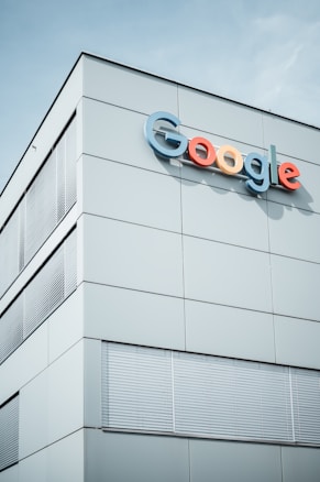 A modern building with the Google logo displayed prominently on the upper section. The structure features minimalist design with large, smooth panels and multiple sets of window blinds. The sky is clear in the background.