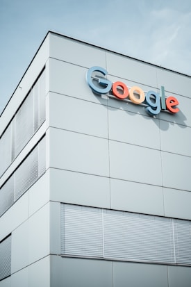 A modern building with the Google logo displayed prominently on the upper section. The structure features minimalist design with large, smooth panels and multiple sets of window blinds. The sky is clear in the background.