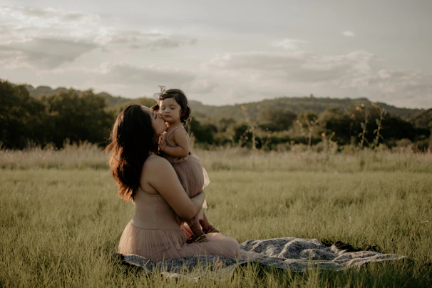 A serene portrait of a mother and child sitting on a terracotta-colored blanket in a sunlit meadow.