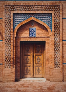 An intricately designed wooden doorway is set within a brick wall adorned with geometric blue tile patterns. The door features carved panels and is framed by an arch accented with blue and white floral tiles.