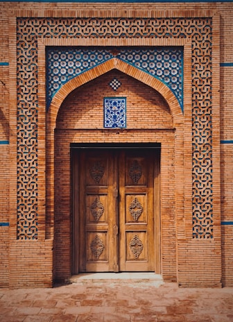 An intricately designed wooden doorway is set within a brick wall adorned with geometric blue tile patterns. The door features carved panels and is framed by an arch accented with blue and white floral tiles.