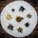 An assortment of various dried tea leaves and blossoms is arranged on a circular white cloth placed on a wooden surface. The teas include dark loose leaf piles, a small group of yellow blossoms, and a round citrus shell filled with tea leaves.