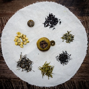 An assortment of various dried tea leaves and blossoms is arranged on a circular white cloth placed on a wooden surface. The teas include dark loose leaf piles, a small group of yellow blossoms, and a round citrus shell filled with tea leaves.