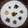 An assortment of various dried tea leaves and blossoms is arranged on a circular white cloth placed on a wooden surface. The teas include dark loose leaf piles, a small group of yellow blossoms, and a round citrus shell filled with tea leaves.
