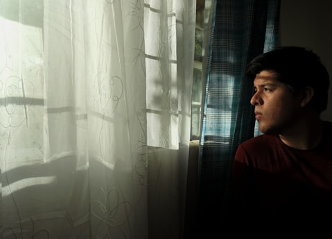 A person is gazing thoughtfully out of a window. Sunlight filters through patterned curtains, casting soft shadows inside the room. The light and shadow create an introspective and serene atmosphere.