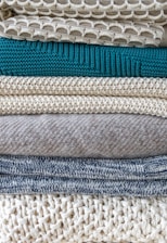 A stack of neatly folded knitted sweaters showcasing a variety of textures and patterns. The pile features different knit designs, including a honeycomb pattern and ribbed styles, in neutral tones with one teal garment in the middle.