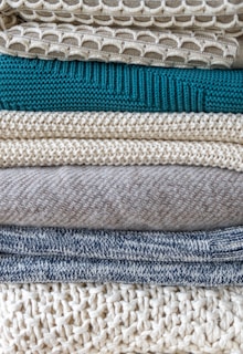 A stack of neatly folded knitted sweaters showcasing a variety of textures and patterns. The pile features different knit designs, including a honeycomb pattern and ribbed styles, in neutral tones with one teal garment in the middle.