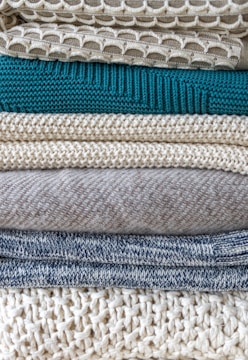 A stack of neatly folded knitted sweaters showcasing a variety of textures and patterns. The pile features different knit designs, including a honeycomb pattern and ribbed styles, in neutral tones with one teal garment in the middle.