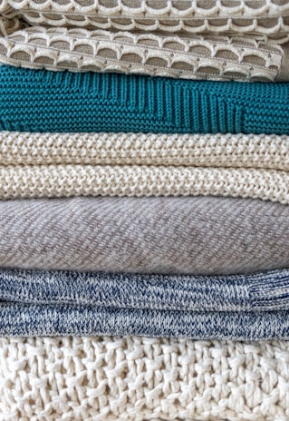 A stack of neatly folded knitted sweaters showcasing a variety of textures and patterns. The pile features different knit designs, including a honeycomb pattern and ribbed styles, in neutral tones with one teal garment in the middle.