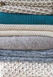 A stack of neatly folded knitted sweaters showcasing a variety of textures and patterns. The pile features different knit designs, including a honeycomb pattern and ribbed styles, in neutral tones with one teal garment in the middle.