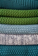 Multiple layers of knitted fabric are neatly stacked, showcasing different textures and patterns. The fibers vary in thickness, giving each layer a distinct tactile quality. The colors transition from deep teal at the top, through shades of green and blue, to a patterned white and navy knit.