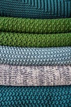 Multiple layers of knitted fabric are neatly stacked, showcasing different textures and patterns. The fibers vary in thickness, giving each layer a distinct tactile quality. The colors transition from deep teal at the top, through shades of green and blue, to a patterned white and navy knit.