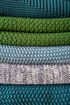 Multiple layers of knitted fabric are neatly stacked, showcasing different textures and patterns. The fibers vary in thickness, giving each layer a distinct tactile quality. The colors transition from deep teal at the top, through shades of green and blue, to a patterned white and navy knit.