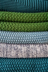Multiple layers of knitted fabric are neatly stacked, showcasing different textures and patterns. The fibers vary in thickness, giving each layer a distinct tactile quality. The colors transition from deep teal at the top, through shades of green and blue, to a patterned white and navy knit.