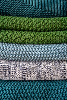 Multiple layers of knitted fabric are neatly stacked, showcasing different textures and patterns. The fibers vary in thickness, giving each layer a distinct tactile quality. The colors transition from deep teal at the top, through shades of green and blue, to a patterned white and navy knit.