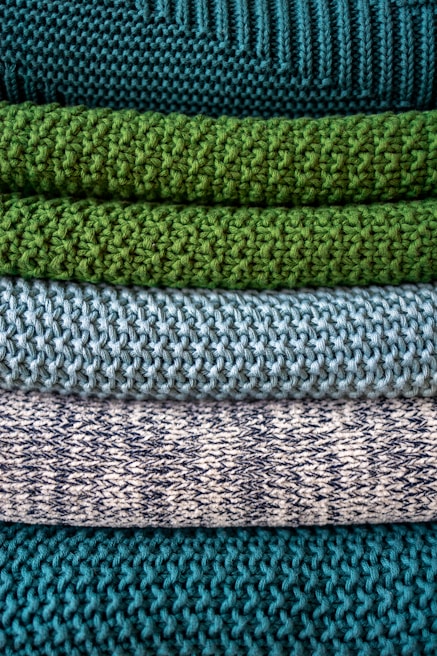 Multiple layers of knitted fabric are neatly stacked, showcasing different textures and patterns. The fibers vary in thickness, giving each layer a distinct tactile quality. The colors transition from deep teal at the top, through shades of green and blue, to a patterned white and navy knit.