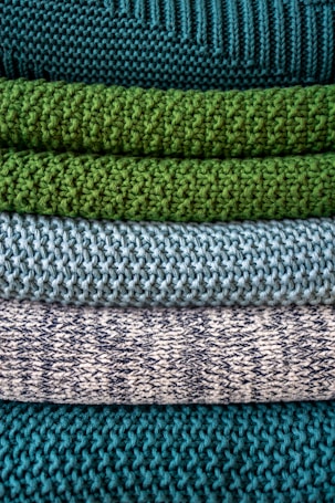 Multiple layers of knitted fabric are neatly stacked, showcasing different textures and patterns. The fibers vary in thickness, giving each layer a distinct tactile quality. The colors transition from deep teal at the top, through shades of green and blue, to a patterned white and navy knit.