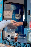 Two hands are engaged in a transaction at an ice cream truck, with one hand receiving change. The truck's interior is visible, showing an ice cream dispenser, a carton box labeled as containing cones, and various stickers on the window. A cup of ice cream with chocolate sauce and a flake is placed on the counter next to a card reader device.