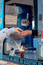 Two hands are engaged in a transaction at an ice cream truck, with one hand receiving change. The truck's interior is visible, showing an ice cream dispenser, a carton box labeled as containing cones, and various stickers on the window. A cup of ice cream with chocolate sauce and a flake is placed on the counter next to a card reader device.