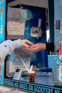 Two hands are engaged in a transaction at an ice cream truck, with one hand receiving change. The truck's interior is visible, showing an ice cream dispenser, a carton box labeled as containing cones, and various stickers on the window. A cup of ice cream with chocolate sauce and a flake is placed on the counter next to a card reader device.